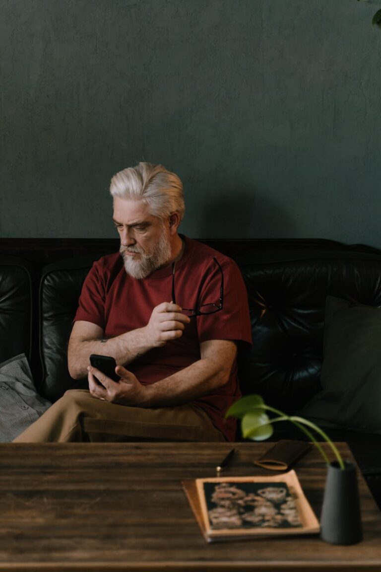 A senior man sits thoughtfully on a sofa, holding a phone and eyeglasses, in a cozy room.