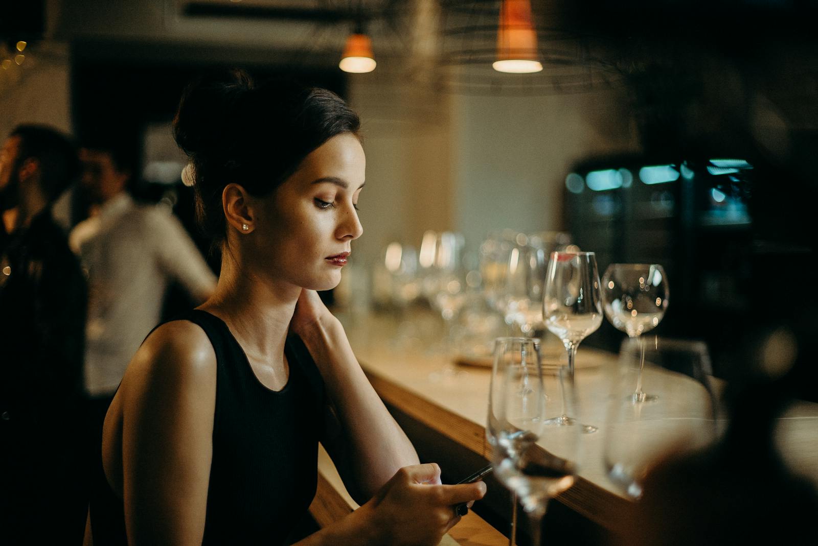 Young woman in a bar setting, elegantly dressed, texting on her phone. Soft lighting enhances the warm atmosphere.