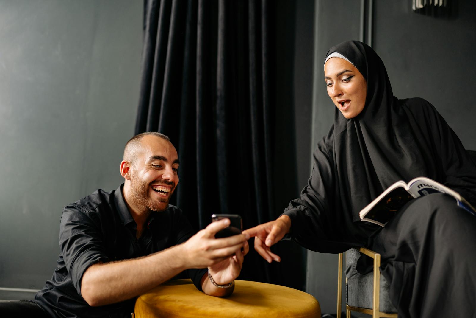 A happy couple in traditional attire laughing and using a smartphone indoors.