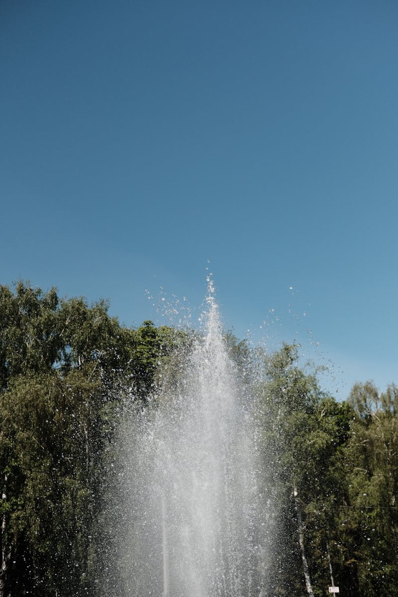A vertical shot of a fountain with water spraying against a clear blue sky in a park.