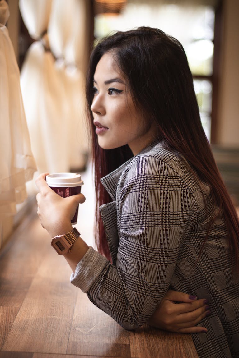 Stylish Asian woman in plaid coat sipping coffee by a sunlit window indoors.