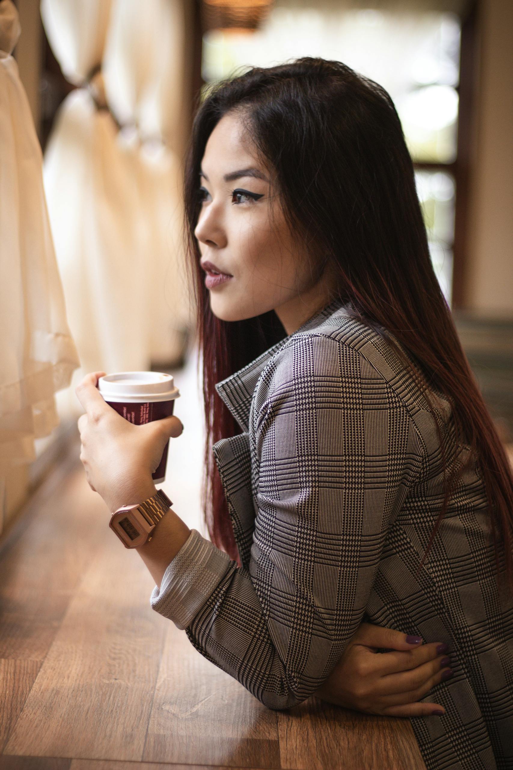 Stylish Asian woman in plaid coat sipping coffee by a sunlit window indoors.