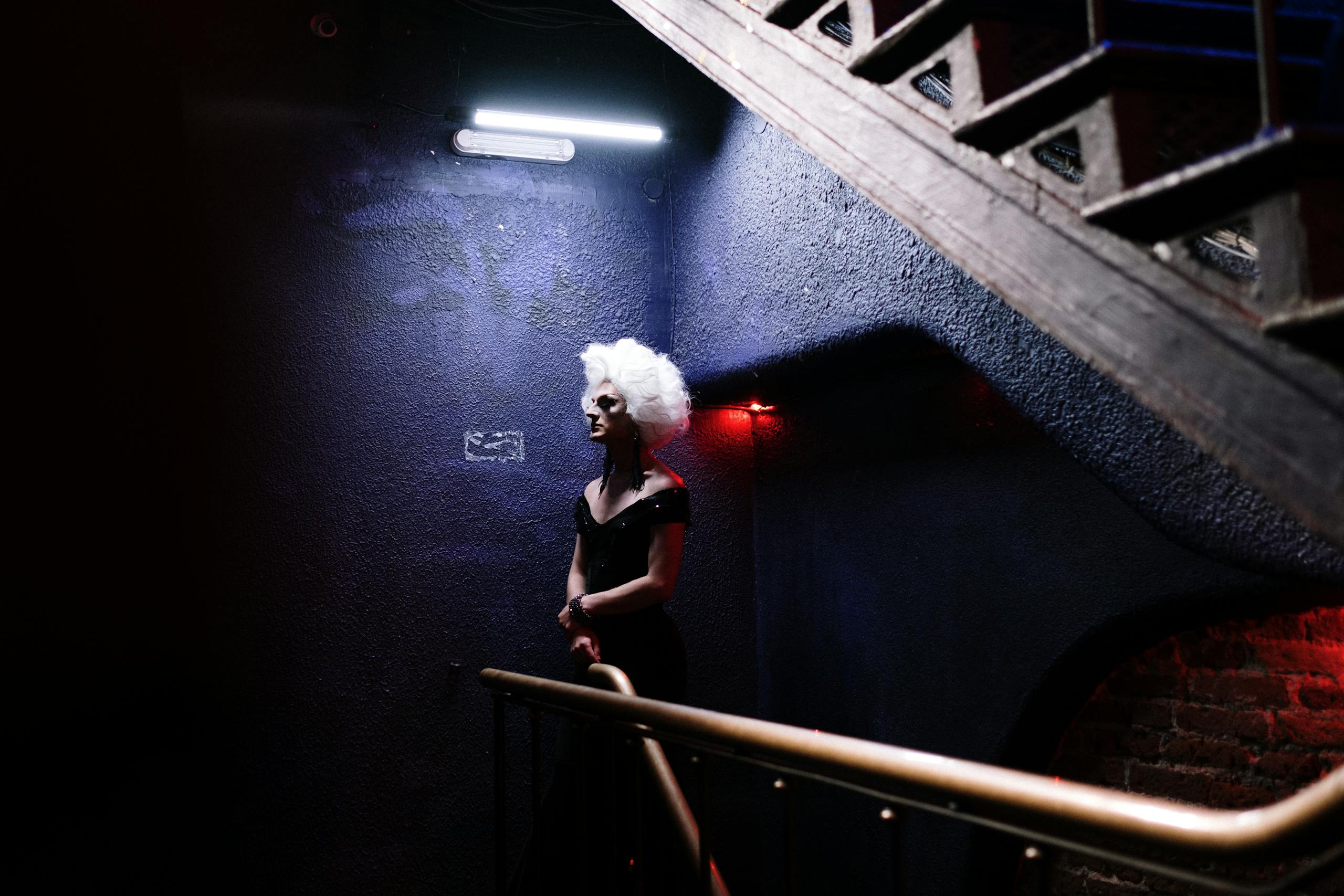 A striking drag queen standing elegantly on a dimly lit staircase in a nightclub setting.