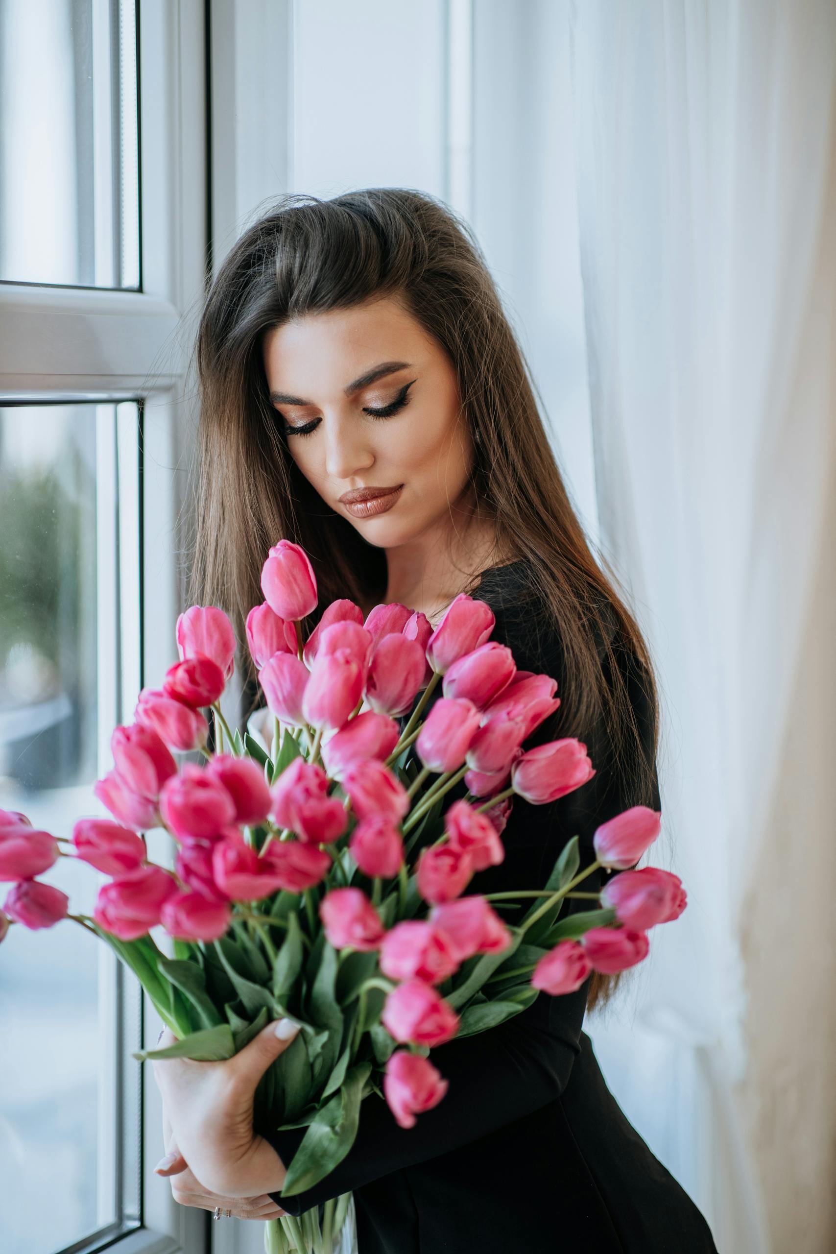 A woman in a black dress holding a bouquet of pink tulips by a bright window indoors.