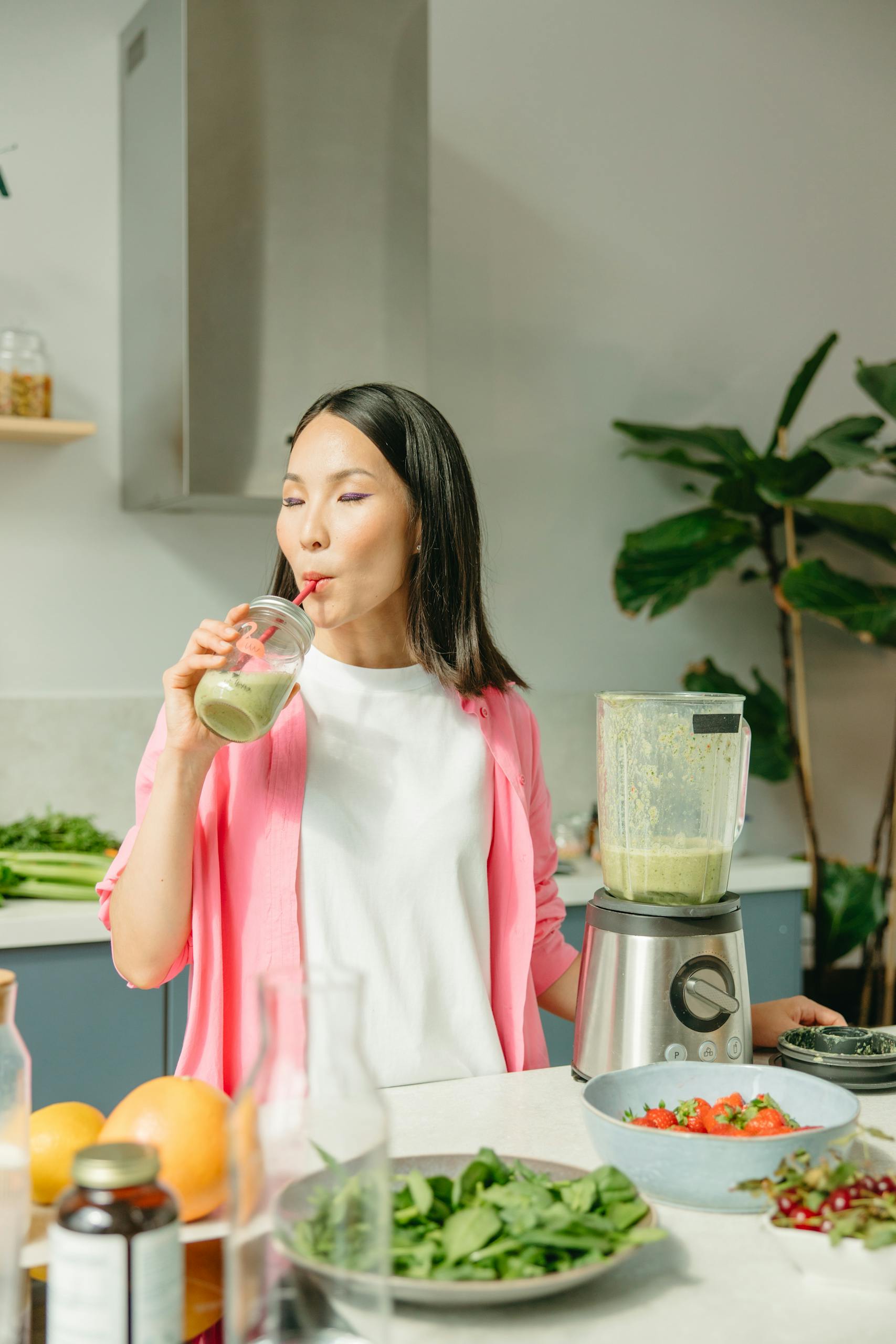 Asian woman sipping a green smoothie in a modern kitchen setting.