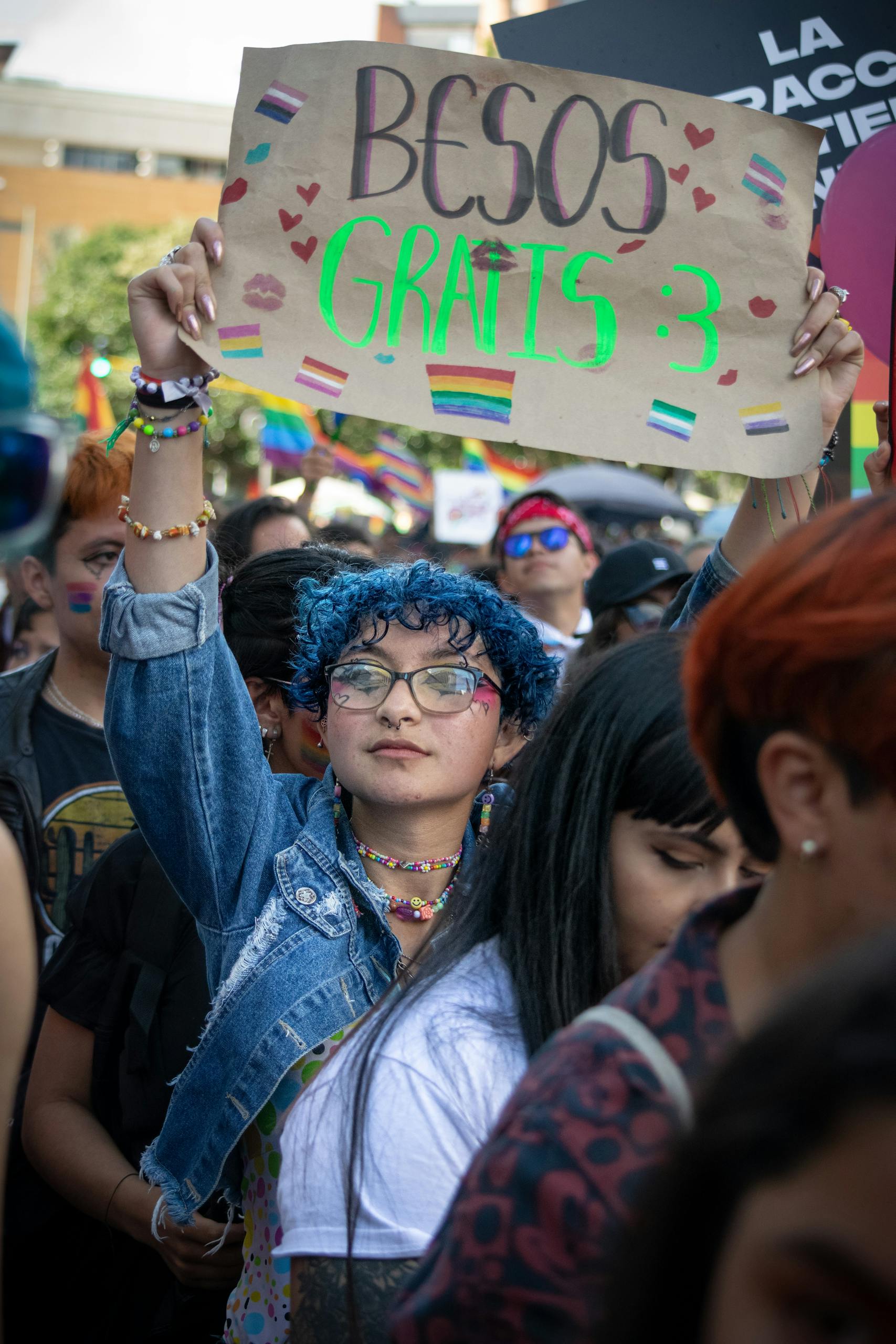 Colorful crowd at Bogotá protest, person holding 'Besos Gratis' sign with raised arms.