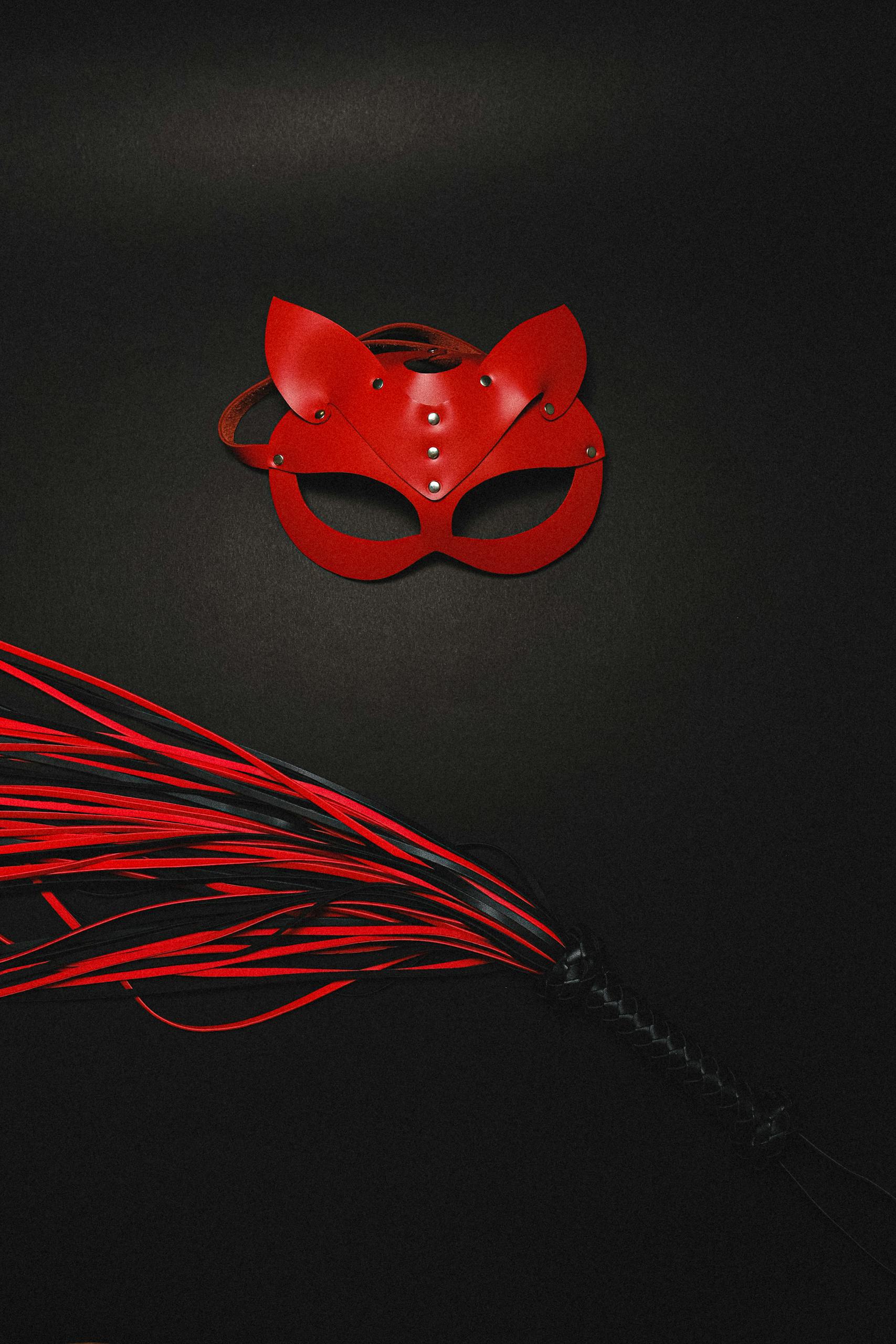 Red leather mask and whip laid on a black surface, symbolizing sensuality and mystery.