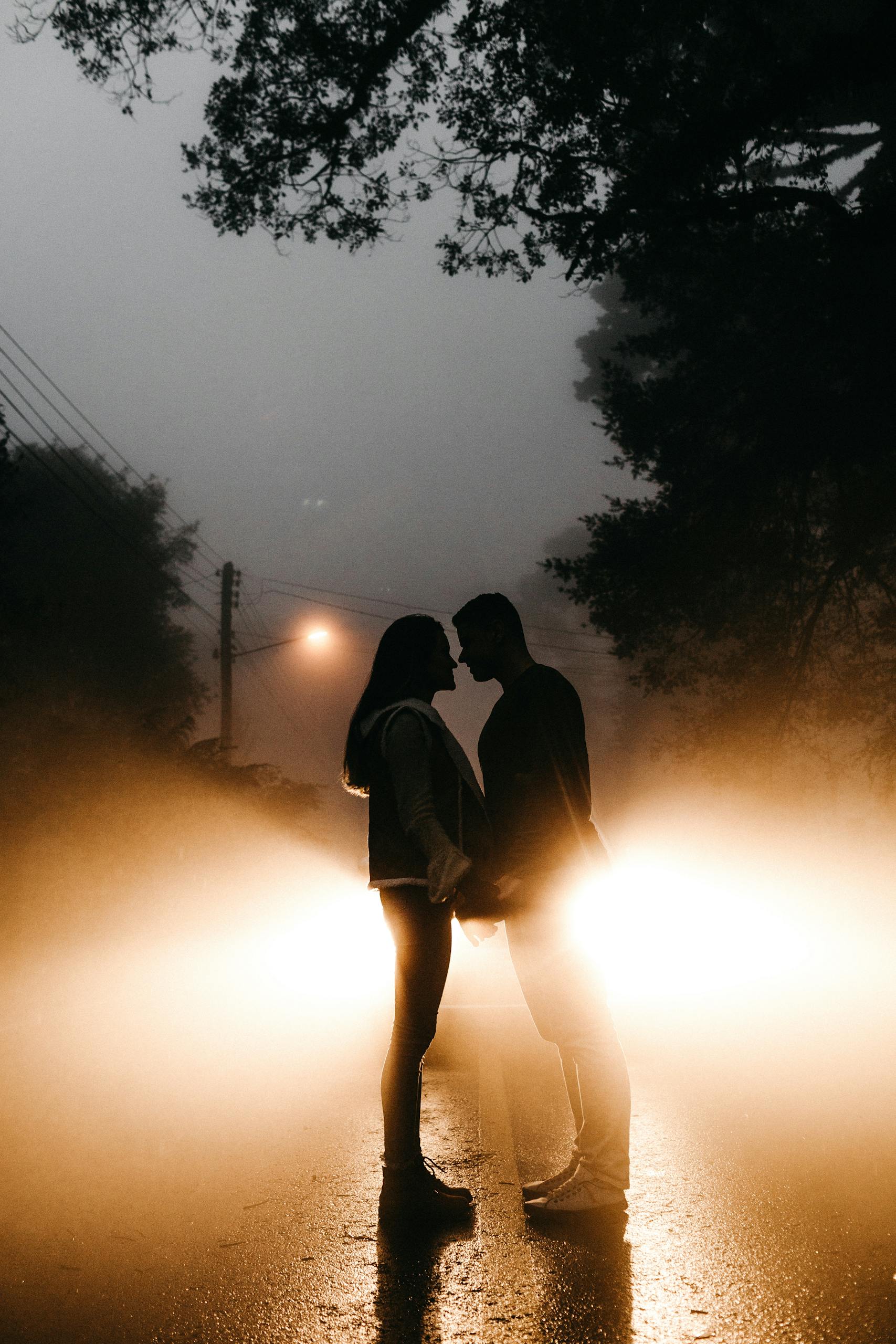 Silhouetted couple standing in front of car headlights on a foggy night, embracing romance.