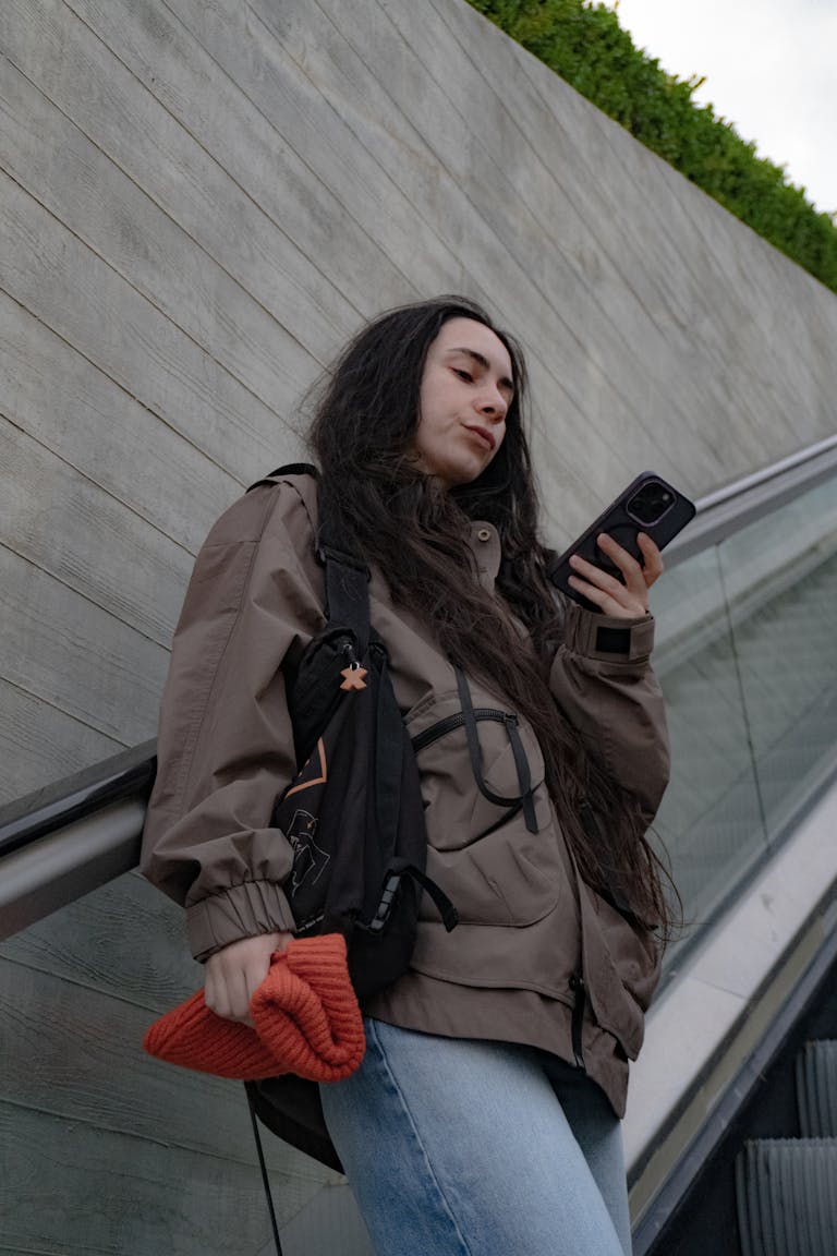 Casual young woman checks her phone while riding an outdoor escalator dressed in a jacket and jeans.